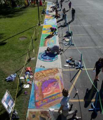 Artists create colorful chalk drawings on the pavement at an outdoor event. The artwork features vibrant images and book covers, with people working and supplies scattered along the sidewalk.