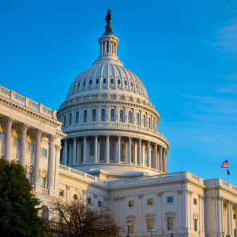 The white dome of the United States Capitol building is shown against a clear blue sky, with part of the American flag visible and sunlight illuminating the structure.