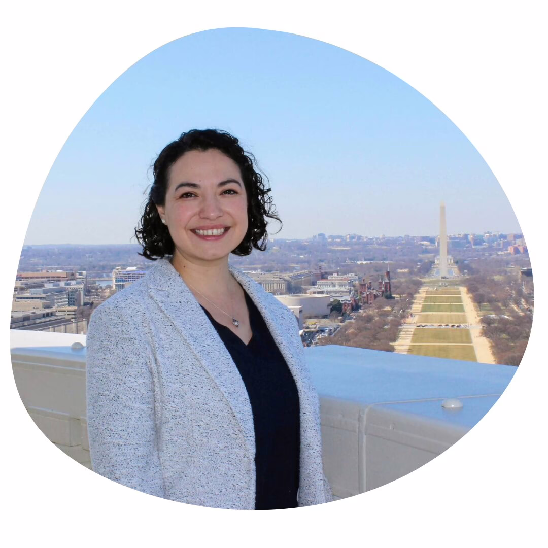 A woman with curly dark hair, wearing a light gray blazer and dark top, smiles while standing on a rooftop with the Washington Monument and National Mall visible in the background on a clear day.