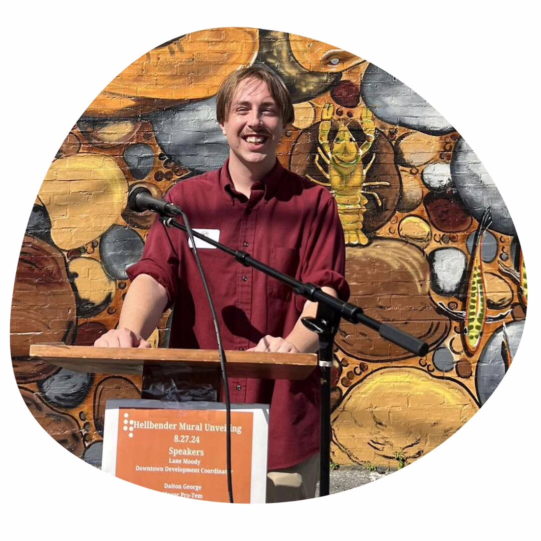 A person in a red shirt stands smiling at a podium with a microphone, in front of a colorful mural featuring a large salamander and other nature-themed artwork.