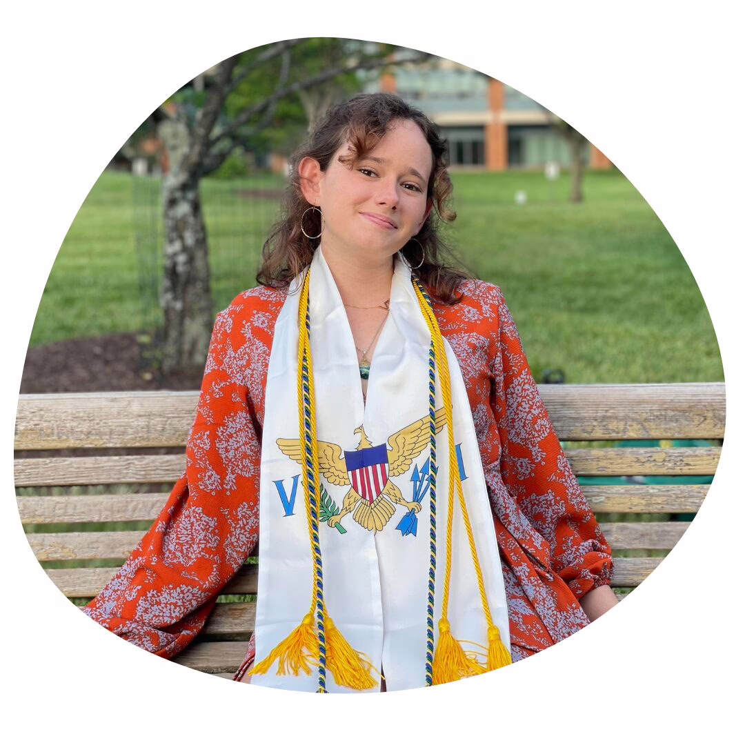 A young woman with curly hair sits on a bench outdoors, smiling. She wears a red patterned jacket and a white graduation stole with an eagle emblem and gold tassels. Grass and trees are visible in the background.