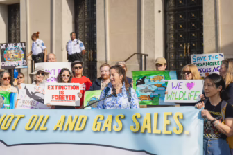 Alt text: A group of wildlife and climate activists stand on building steps behind a speaker at a microphone, holding colorful signs reading “Extinction Is Forever,” “We ♥ Wildlife,” and “Save the Endangered Species Act,” while a large banner in front says “Not Oil and Gas Sales.”