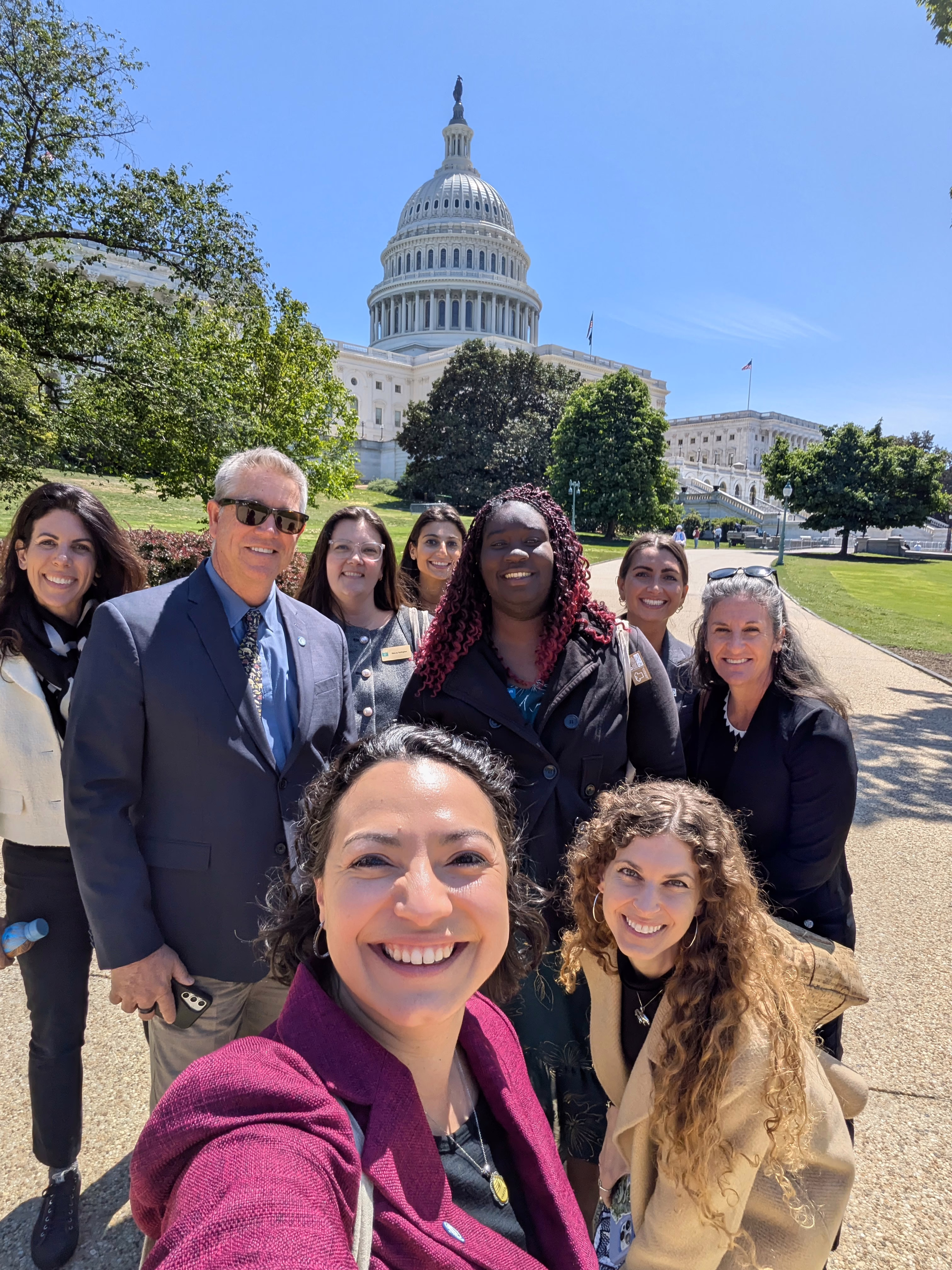 Group selfie of eight smiling people standing on the Capitol grounds in Washington, D.C., with the U.S. Capitol dome rising behind them under a clear blue sky.