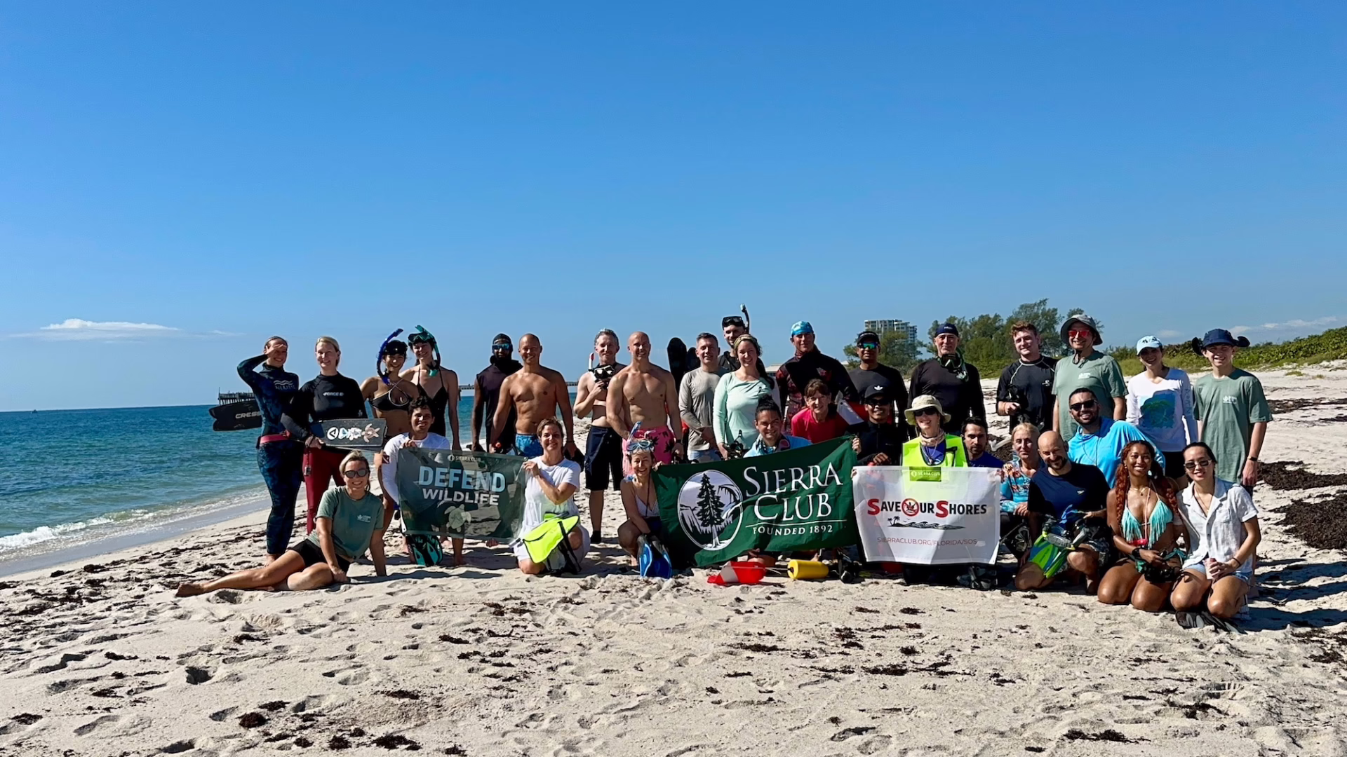 A group of volunteers poses on a sandy beach after a coastal cleanup, holding banners for Defend Wildlife, Sierra Club, and Save Our Shores.