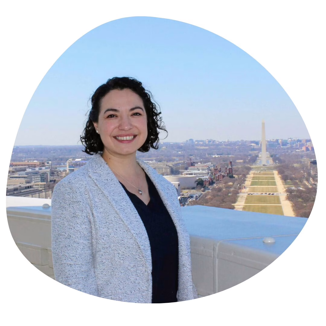 A woman with curly dark hair, wearing a light gray blazer and dark top, smiles while standing on a rooftop with the Washington Monument and National Mall visible in the background on a clear day.