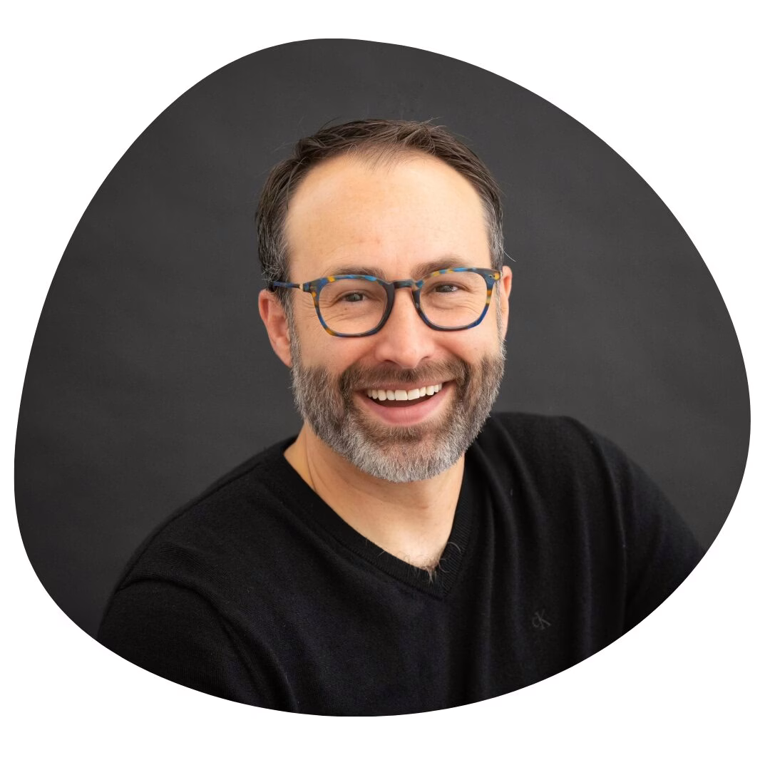 A man with short brown hair, glasses, and a beard smiles at the camera. He is wearing a black shirt and is posed in front of a dark, rounded background.